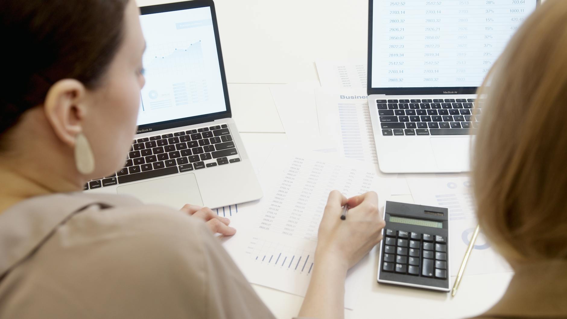 Ilustrasi: Two people analyzing business data on laptops with charts and graphs.