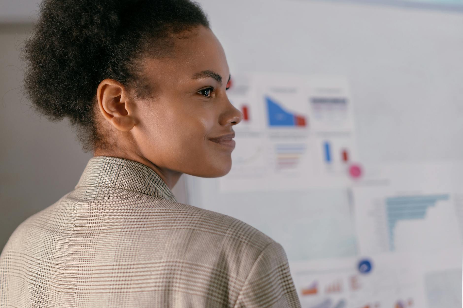 Ilustrasi: A confident businesswoman with curly hair smiling while presenting data charts in an office.